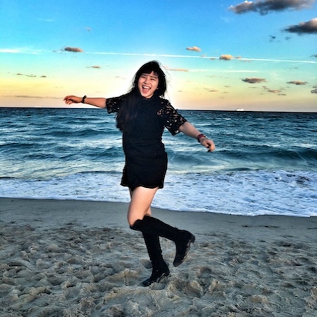 Zhen Cao in black dree and black knee high boots on the sand with ocean in bachground